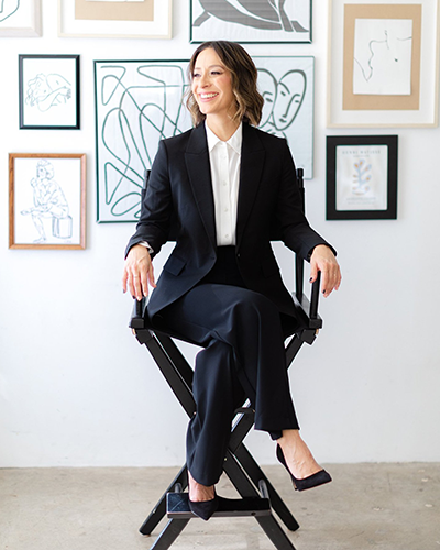 A full-length shot of Olga Brandeis sitting in a director's chair in front of a gallery wall with black and white, framed, drawings.